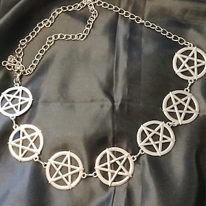 May include: Silver-coloured necklace with circular pendants, each displaying a five-pointed star. The necklace is set against a dark, textured backdrop, highlighting the bright metal of the chain and pendants.