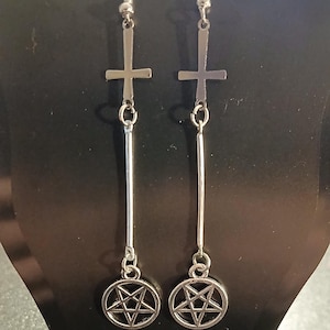May include: Silver-toned dangle earrings. Each earring features a cross at the top, a vertical bar, and a pentagram charm. The earrings are displayed against a black background.