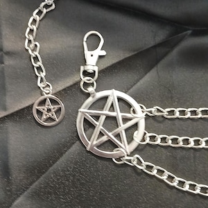 May include: Silver-toned metal chain belt featuring a pentagram pendant and smaller charm. The belt has multiple chain strands and a clasp closure. The design is set against a dark background, highlighting the metallic shine of the accessory.