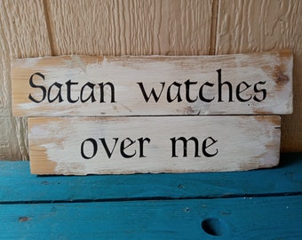 May include: Two wooden planks with the text "Satan watches over me" in black lettering. The planks have a distressed, rustic look with a white painted background and visible wood grain.