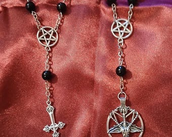 May include: Two rosary-style necklaces with black beads and silver-tone metal accents. One features a cross pendant, the other a pentagram with a goat head. The background is a deep red fabric.