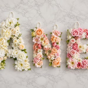 Könnte beinhalten: Drei Wandbuchstaben, die A, N und E bilden und aus künstlichen Blumen bestehen. Der Buchstabe A zeigt weiße und cremefarbene Blüten, während N und E mit einer Mischung aus pfirsichfarbenen, korallenroten und rosa Blumen verziert sind. Jeder Buchstabe hat eine Perlenschlaufe.