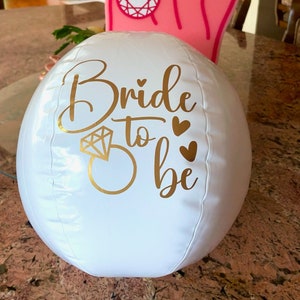 Peut inclure: Ballon de plage gonflable blanc avec l'inscription dorée "Bride to be" avec un graphique de bague en diamant et trois petits cœurs.