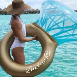 Pode incluir: Um flutuador inflável dourado em forma de anel com um topo azul em forma de diamante. As palavras "Bride to Be" estão escritas em branco na lateral do anel. O flutuador é segurado por uma pessoa vestindo um maiô branco e um chapéu de palha.