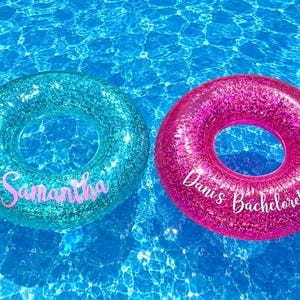 Könnte beinhalten: Zwei kreisförmige aufblasbare Schwimmringe mit glitzernden Oberflächen. Ein Schwimmring ist türkis mit dem Namen Samantha in rosa Schreibschrift. Der andere Schwimmring ist pink mit dem Text Dani's Bachelorette in weißer Schrift.