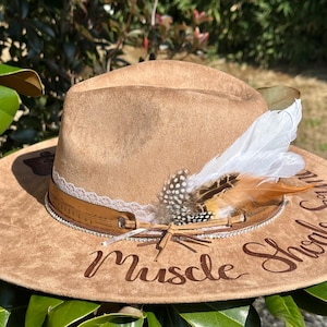 Op de afbeelding: Een bruine su&egrave;de cowboyhoed met een witte en bruine verenversiering. De hoed heeft een kanten rand en een bruin leren band met een muzieknootontwerp. De woorden "Muscle Shoals" zijn in bruin op de hoed geborduurd.