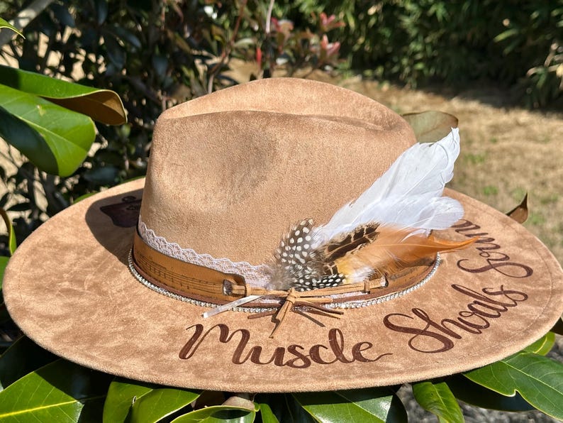 Op de afbeelding: Een bruine su&egrave;de cowboyhoed met een witte en bruine verenversiering. De hoed heeft een kanten rand en een bruin leren band met een muzieknootontwerp. De woorden "Muscle Shoals" zijn in bruin op de hoed geborduurd.
