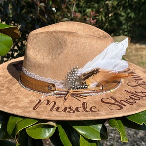 Op de afbeelding: Een bruine su&egrave;de cowboyhoed met een witte en bruine verenaccent. De hoed heeft een bruin leren band met strass steentjes en een witte kantversiering. De woorden "Muscle Shoals Sound" zijn in bruin schrift op de hoed geschreven.
