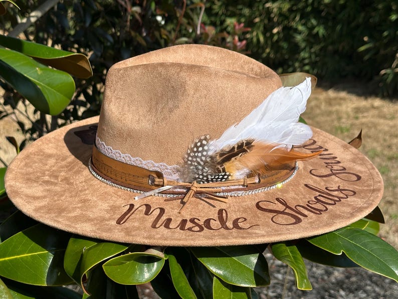 Op de afbeelding: Een bruine su&egrave;de cowboyhoed met een witte en bruine verenaccent. De hoed heeft een bruin leren band met strass steentjes en een witte kantversiering. De woorden "Muscle Shoals Sound" zijn in bruin schrift op de hoed geschreven.