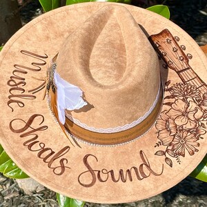 Op de afbeelding: Een bruine fedora hoed van vilt met een bruine leren band en een witte kantversiering. De hoed heeft een bloemenmotief en een gitaar aan de zijkant. De tekst "Hustle Shawls Sound" staat op de hoed.