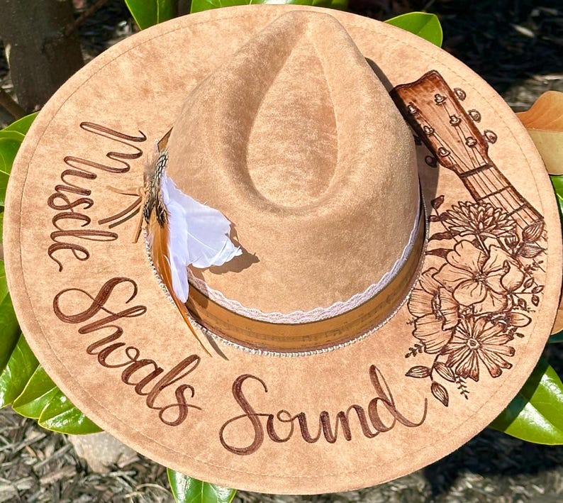 Op de afbeelding: Een bruine fedora hoed van vilt met een bruine leren band en een witte kantversiering. De hoed heeft een bloemenmotief en een gitaar aan de zijkant. De tekst "Hustle Shawls Sound" staat op de hoed.