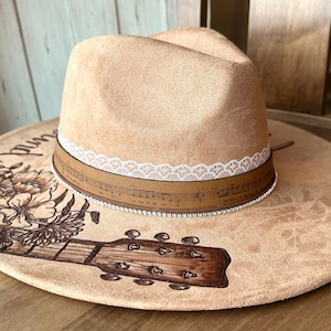 Op de afbeelding: Een lichtbruine wollen fedora-hoed met een witte kantversiering en een bruin leren band met muzieknoten. De hoed heeft een gitaar- en bloemenontwerp op de rand.