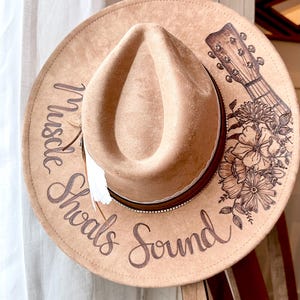 Op de afbeelding: Een bruine cowboyhoed van vilt met een leren band en een witte veer. De hoed is versierd met een bloemenpatroon en een gitaar. De tekst "Muscle Shoals Sound" staat op de hoed.