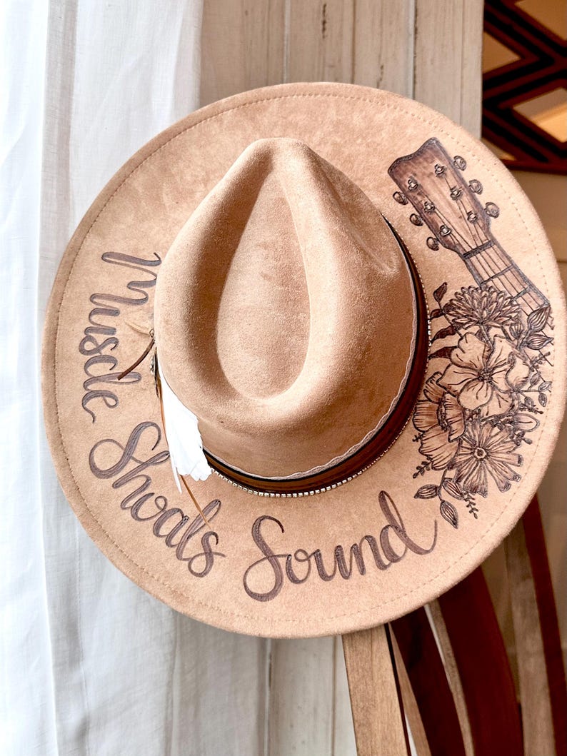 Op de afbeelding: Een bruine cowboyhoed van vilt met een leren band en een witte veer. De hoed is versierd met een bloemenpatroon en een gitaar. De tekst "Muscle Shoals Sound" staat op de hoed.