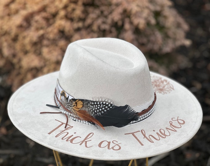 Lainey Wilson Cowboy Hat with Thick as Thieves wording and burned flowers on the brim, lace and feather accents with ticket stub