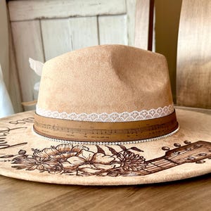 Op de afbeelding: Een bruine su&egrave;de cowboyhoed met een witte kanten rand en een bruin leren band met muzieknoten. De hoed is versierd met een bloemenontwerp en een gitaar. De rand van de hoed heeft de woorden "Around the" erop geschreven.
