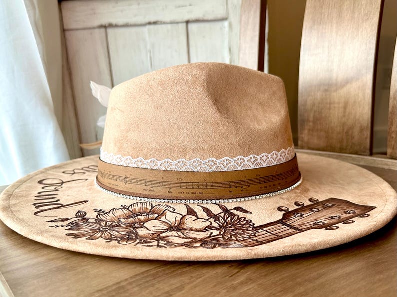 Op de afbeelding: Een bruine su&egrave;de cowboyhoed met een witte kanten rand en een bruin leren band met muzieknoten. De hoed is versierd met een bloemenontwerp en een gitaar. De rand van de hoed heeft de woorden "Around the" erop geschreven.