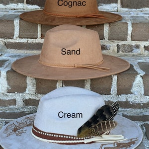 Op de afbeelding: Drie vilten cowboyhoeden op elkaar gestapeld. De bovenste hoed is bruin en heeft de tekst "Cognac". De middelste hoed is beige en heeft de tekst "Sand". De onderste hoed is cr&egrave;mekleurig met een bruine band en een veer en heeft de tekst "Cream".