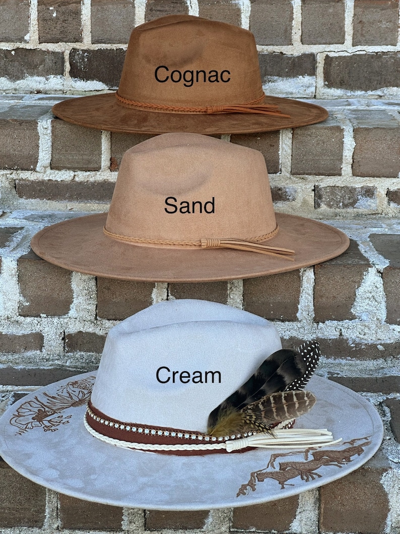 Op de afbeelding: Drie vilten cowboyhoeden op elkaar gestapeld. De bovenste hoed is bruin en heeft de tekst "Cognac". De middelste hoed is beige en heeft de tekst "Sand". De onderste hoed is cr&egrave;mekleurig met een bruine band en een veer en heeft de tekst "Cream".