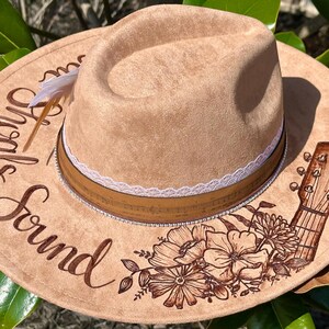 Op de afbeelding: Een beige su&egrave;de fedora-hoed met een witte kanten band en een bruine leren band. De hoed is versierd met een bloemenontwerp en een gitaar. De tekst "Sounds Good" staat op de hoed.
