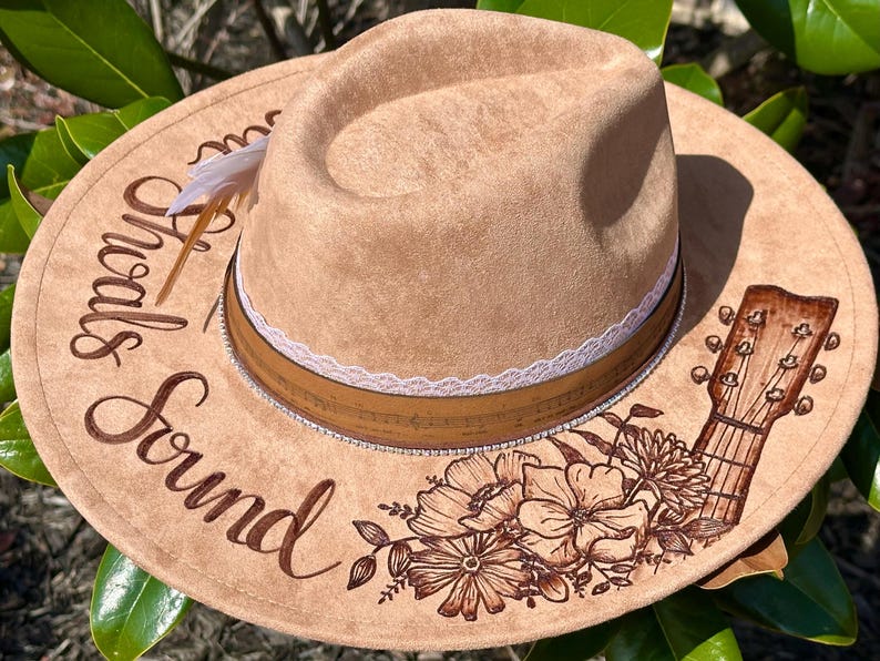 Op de afbeelding: Een beige su&egrave;de fedora-hoed met een witte kanten band en een bruine leren band. De hoed is versierd met een bloemenontwerp en een gitaar. De tekst "Sounds Good" staat op de hoed.