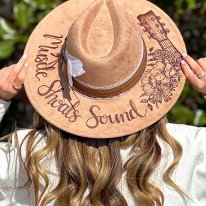 Op de afbeelding: Een bruine fedora hoed van vilt met een witte kantband en een bloemenmotief. De hoed heeft de woorden "Hustle" en "Sounds" erop geschreven, samen met een gitaargrafiek.