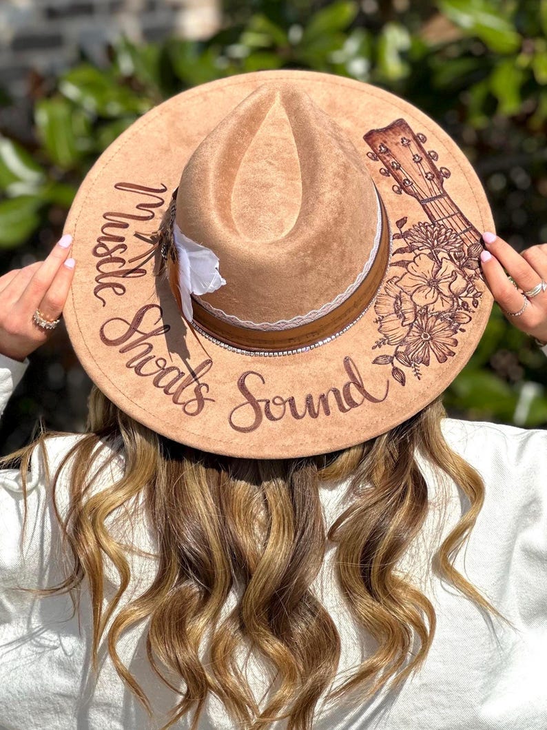 Op de afbeelding: Een bruine fedora hoed van vilt met een witte kantband en een bloemenmotief. De hoed heeft de woorden "Hustle" en "Sounds" erop geschreven, samen met een gitaargrafiek.