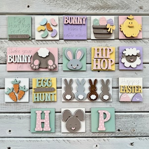 May include: A set of 12 wooden Easter signs with various designs, including bunnies, eggs, carrots, and the words "Egg Hunting Season Now Open", "Shake Your Bunny Tail", "Egg This Way Hunt", "Bunny Kisses & Easter Wishes", "Hip Hop", "Happy Easter", and "H", "P". The signs are painted in pastel colors and have a rustic, farmhouse style.