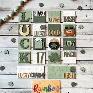 May include: A St. Patrick's Day themed display featuring green and white wooden blocks with phrases like "Lucky Charm" and "Pot of Gold." The blocks are arranged on a white wooden surface, with shamrock cutouts and a beaded garland.