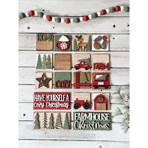 May include: A collection of Christmas-themed wooden blocks with various designs and text, including a mug, deer, car, and the phrase "Farmhouse Christmas." The blocks are arranged on a white wooden background, with a beaded garland above.