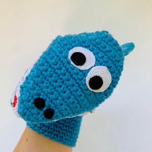 May include: A blue crocheted shark puppet with black eyes and white accents. The puppet is on a hand, with the mouth open and showing white teeth.