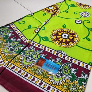 May include: Folded green fabric with a floral pattern and paisley border. The fabric features large yellow and white flowers with maroon and green accents. The border is maroon, yellow, and white with paisley designs. A small blue tag is visible.