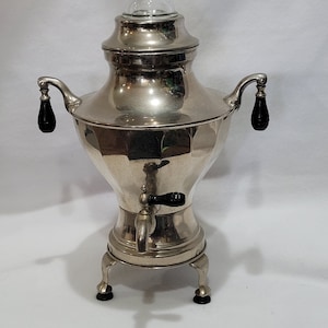 May include: A silver-plated coffee percolator with a glass lid and black plastic handles. The percolator has a decorative design and sits on three legs.