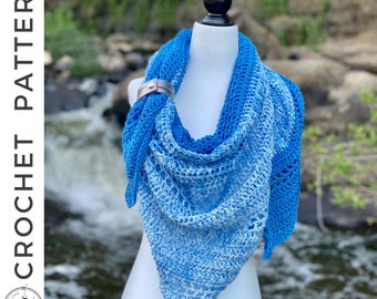 May include: A blue crochet shawl with a gradient effect, transitioning from a deep blue to a lighter shade. The shawl is secured with a silver-toned clasp. The text "CROCHET PATTERN" is visible on the left side.