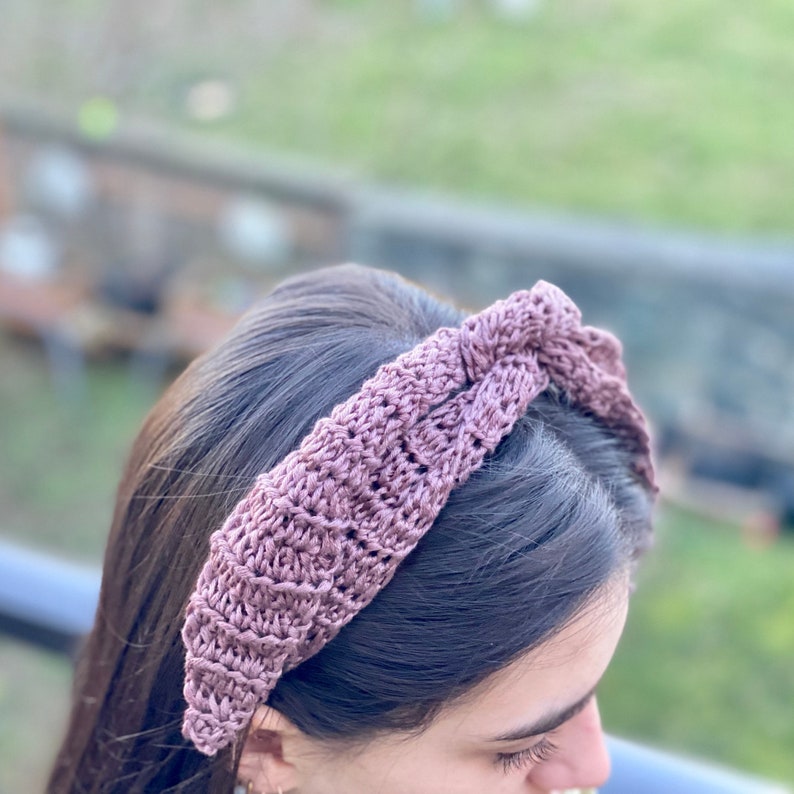 May include: A mauve crocheted headband with a knotted design. The headband is worn on a person's head.