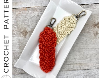 May include: Two crocheted keychains, one orange and one cream, with metal key rings. The keychains are displayed on a white rectangular plate.