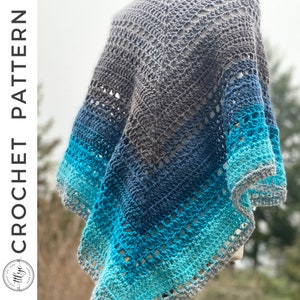 May include: A crocheted shawl in shades of blue and grey, with a lacy pattern. The shawl is draped over a white surface.