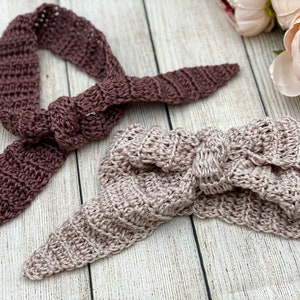 May include: Two crocheted headbands, one in a deep burgundy colour and the other in a light blush pink. Both headbands are tied in a knot and have a textured, woven design.