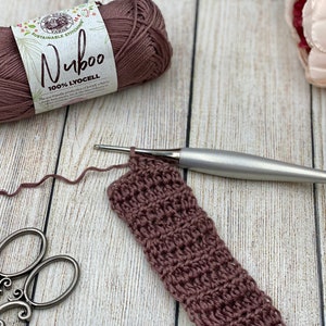 May include: A close-up of a crocheted project in progress, using a maroon yarn and a silver crochet hook. The ball of yarn is in the background, with the label "Nuboo 100% Lyocell" visible.