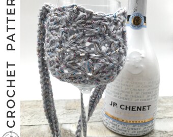 May include: A gray crochet wine glass holder with a strap, featuring metallic thread accents. The holder is wrapped around a clear wine glass, with a bottle of JP Chenet sparkling wine in the background. The words "CROCHET PATTERN" are visible.