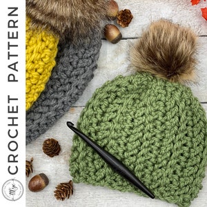 Puede incluir: Un gorro de punto verde con un pompón de piel sintética marrón. El gorro está hecho con un punto texturizado y está sobre una superficie de madera blanca con un gancho de ganchillo.