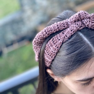 May include: A mauve crocheted headband with a large bow on top. The headband is worn on a person's head.
