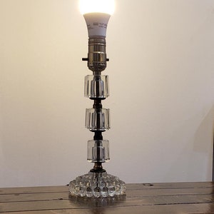 May include: A vintage table lamp featuring a clear glass base and a stacked, cylindrical glass body. The lamp has a metal fixture and a bright, white LED light bulb. The lamp is on a wooden surface.