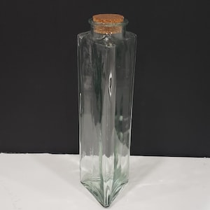 May include: A clear, rectangular glass bottle with a cork stopper. The bottle has an angular design and is empty. The cork is light brown. The bottle is approximately 20 cm tall.