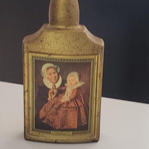 May include: A vintage, rectangular bottle with a dark brown stopper. The bottle is gold-colored with a textured surface and features a framed portrait of two people. The portrait is in a gold frame.