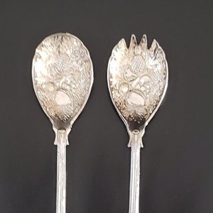 Könnte beinhalten: Ein Set aus zwei silbernen Salatbesteck mit verzierten Griffen. Das Besteck hat ein dekoratives Design auf der Vorder- und Rückseite.