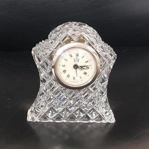 Puede incluir: Un reloj de cristal con una esfera blanca y marcadores de hora romanos negros. El reloj está colocado sobre una base de cristal transparente con un patrón de diamante.