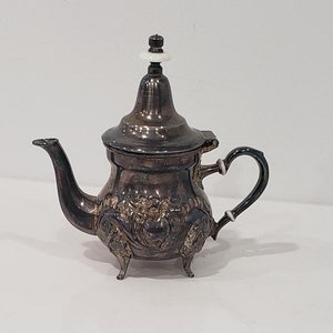 Peut inclure: Une théière en argent avec un design orné et un bouton blanc sur le couvercle. La théière a un bec verseur et une poignée.