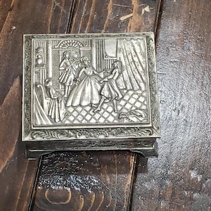 May include: A silver metal trinket box with an ornate design. The box features an embossed scene of four people in a room with a window and a door. The box has a hinged lid and a decorative base.