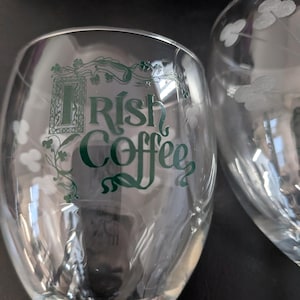 May include: Two clear Irish coffee glasses. One glass has the words "Irish Coffee" in green script. The other glass has a shamrock design etched into the glass. Both glasses are on a dark surface.